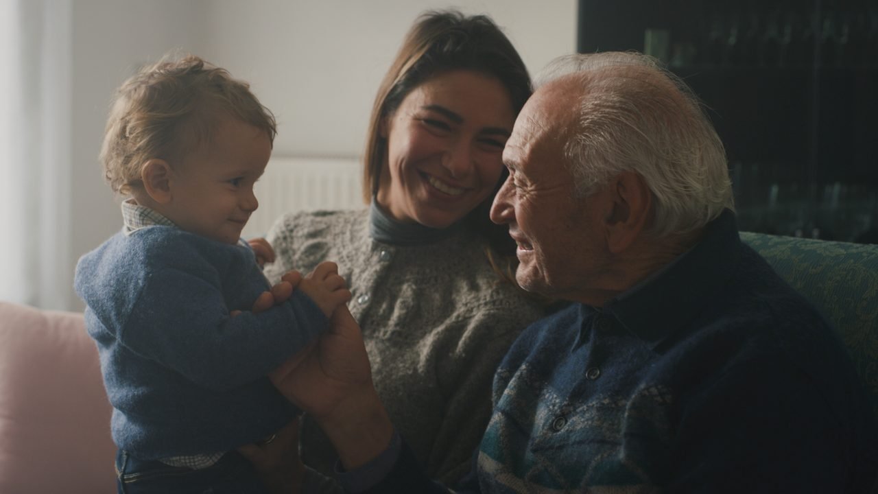 A grandfather, his daughter and her baby laughing together on a sofa.